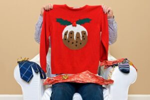 A man opening Christmas presents to discover he got a Christmas themed jumper to go along with the usual socks and tie.